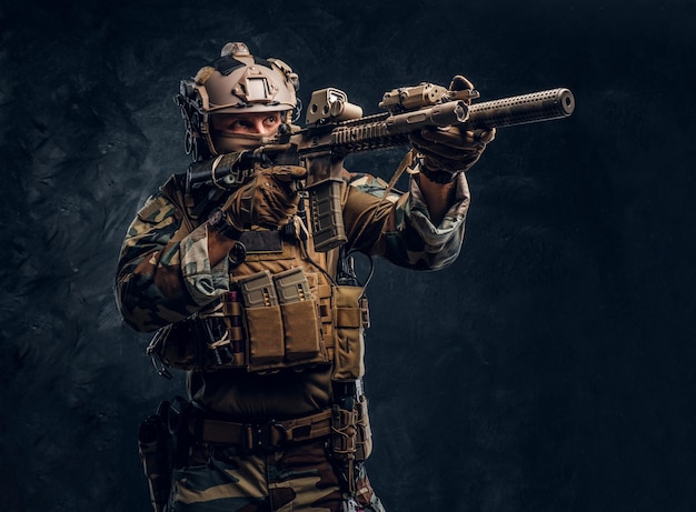 엘리트 부대, 위장 제복을 입은 특수 부대 군인이 돌격 소총을 들고 광학 시력으로 조준합니다. 어두운 질감의 벽에 대한 스튜디오 사진