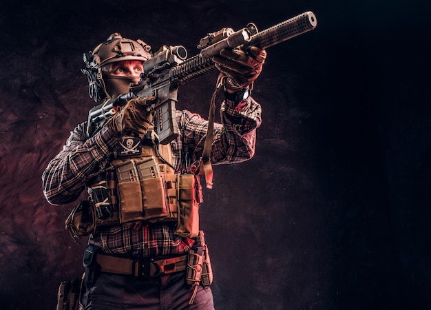 Elite unit, special forces soldier in camouflage uniform holding an assault rifle with a laser sight and aims at the target. Studio photo against a dark textured wall