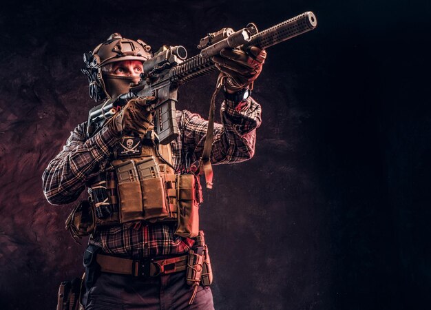 Elite unit, special forces soldier in camouflage uniform holding an assault rifle with a laser sight and aims at the target. Studio photo against a dark textured wall