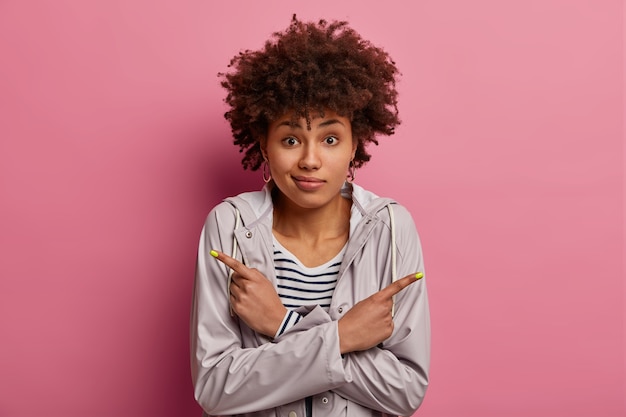 Embarrassed Afro American woman has to make difficult choice, points left and right, looks with questioned hesitant expression, shows two ways, wears grey anorak, isolated on pink wall