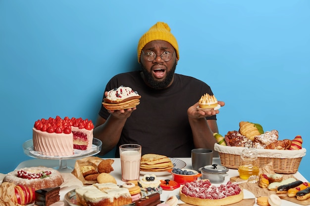 Embarrassed black fat bearded man holds two tasty cakes, cannot make choice what to eat, has tasty sweet breakfast, dressed in casual wear, isolated on blue wall.