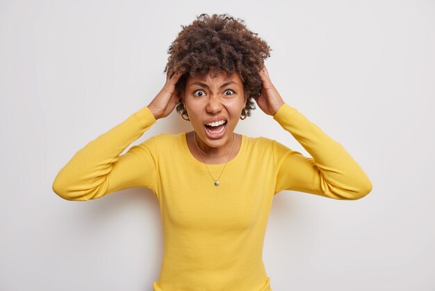 Embarrassed curly haired woman grabs head exclaims loudly feels irritated doesnt know how to solve difficult situation wears yellow sweater isolated over white background Negative emotions