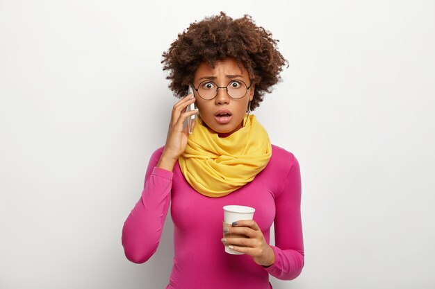 Embarrassed curly woman speaks via smartphone, hears bad information, makes phone call, holds takeout coffee, looks through optical glasses, poloneck and scarf, isolated over white background