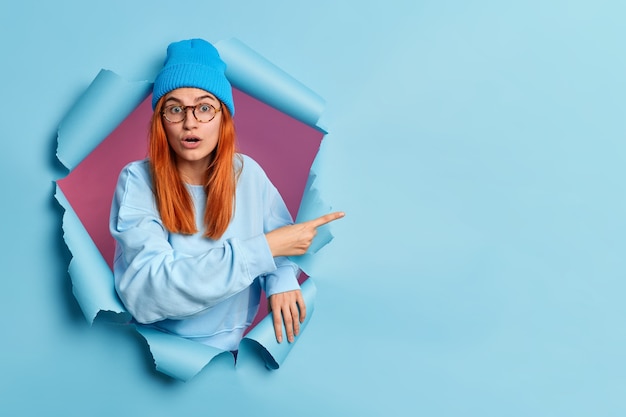 Foto gratuita la donna dai capelli rossi emotiva imbarazzata sta con la bocca aperta, gli occhi infastiditi indica lo spazio della copia attraverso il foro della carta