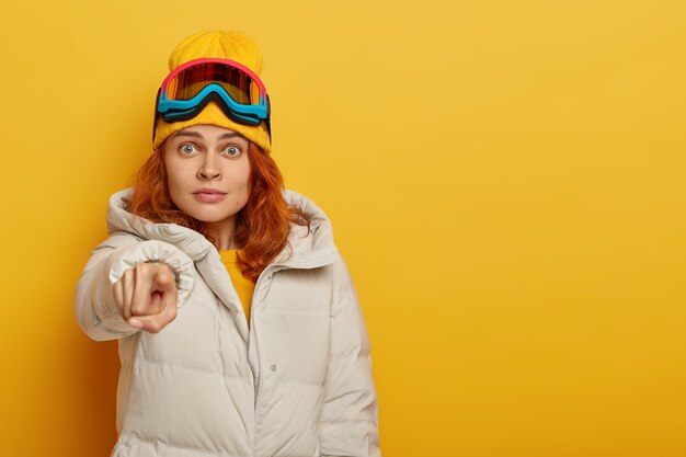 恥ずかしい生姜の女性がカメラを直接指さし、驚きの表情をし、暖かい服を着て、スキーリゾートにいて、冬のツアーをし、黄色の背景と空きスペースで隔離されています