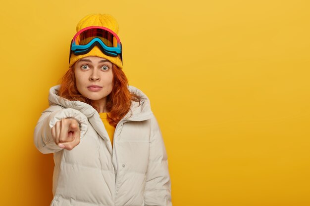 Embarrassed ginger woman points directly at camera, has surprised face expression, dressed in warm clothing, being on ski resort, has winter tour, isolated over yellow background with free space