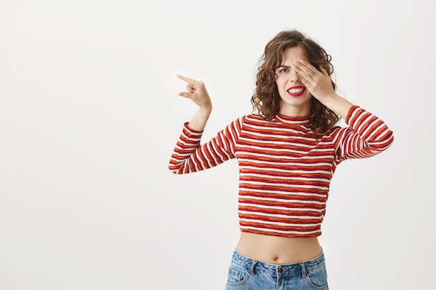 Free photo embarrassed girl closing her eyes and showing something small with disappointed grimace