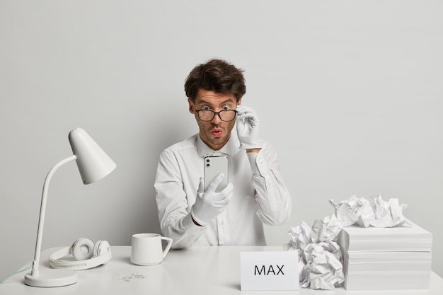 Free photo embarrassed guy stares with wonder at smartphone device, wears transparent glasses