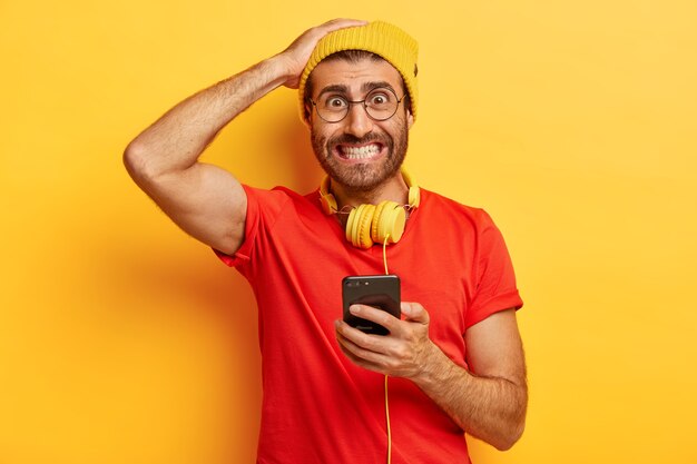 Embarrassed hipster clenches teeth, looks nervously, cannot download necessary application on smart phone, has headphones around neck, dressed casually