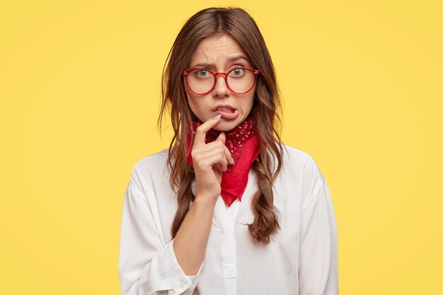 Embarrassed hipster or female teenager has displeased facial expression, keeps index finger near lips, looks with frustration