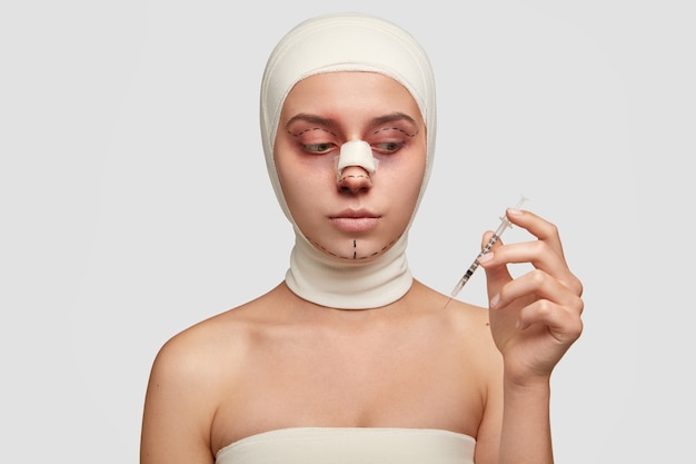 Смущенная модель после ринопластики или изменения формы носа, собирающаяся сделать ритидэктомию, держит шприц с обезболивающим, хочет иметь идеальную мягкую здоровую кожу, изолированную на белом фоне