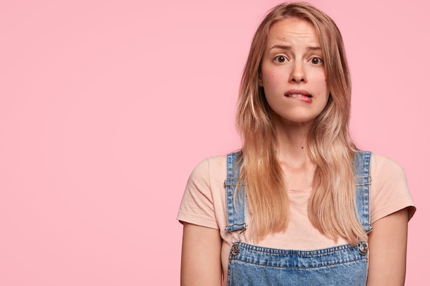 Embarrassed puzzled lovely woman bites lips, has miserable facial expression, dressed in fashionable denim overalls