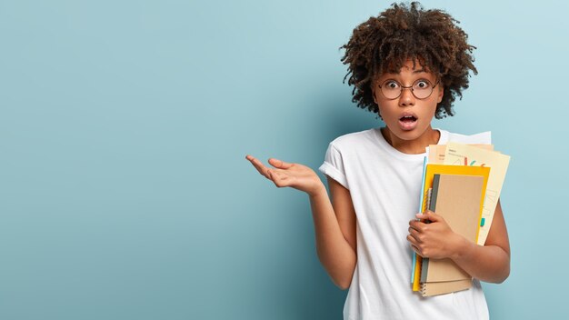 Embarrassed student with Afro appearance, raises palm with bewilderment, wears casual t shirt, holds notepad and papers, prepares for examination, puzzled by much material to learn, has deadline