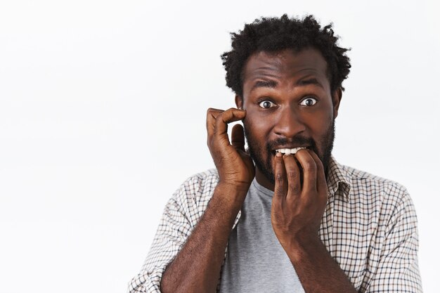 Embarrassed and worried bearded african-american guy feel panic, biting nails and scratch ear, stare anxious