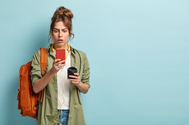 Смущенный молодой студент колледжа с причесанными волосами, в повседневной рубашке, удивленный плохим подключением к Интернету, смотрит на экран мобильного телефона, загружает музыку из плейлиста, пьет кофе на вынос