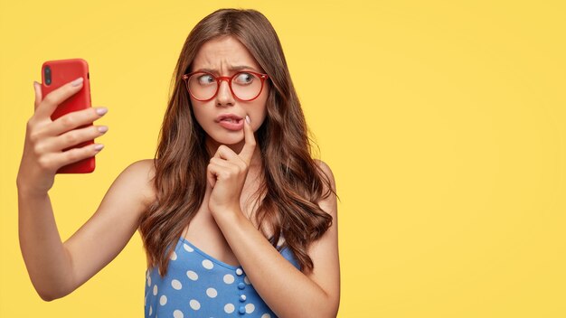 Embarrassed young European lady with nervous worried expression, wears spectacles, holds modern smart phone in front