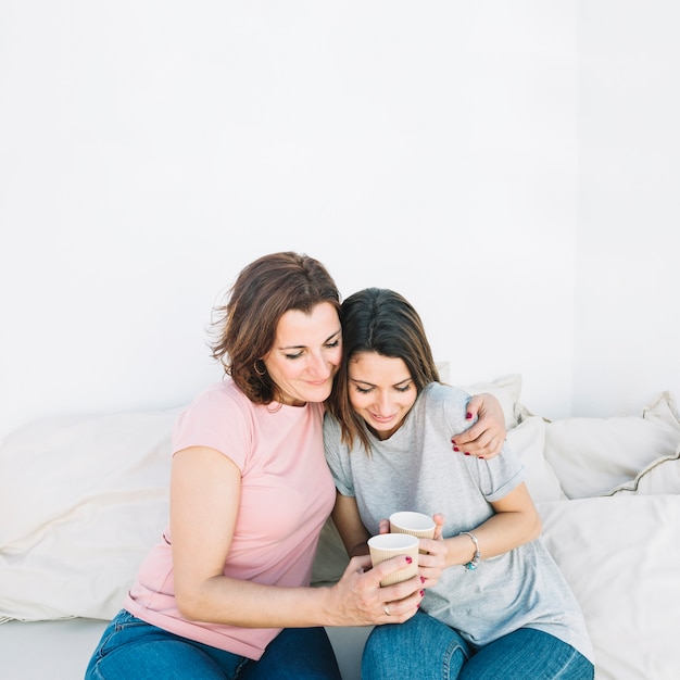 Embracing women with hot beverages on sofa