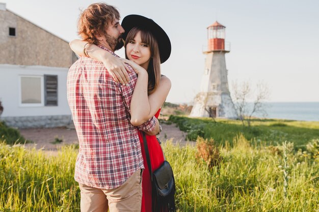 Embracing young stylish couple in love in countryside, indie hipster bohemian style, weekend vacation, summer outfit, red dress, green grass, holding hands