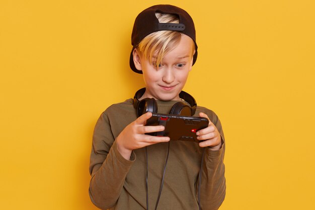 Emotional and active little boy with blond hair, carrying his finger on screen of smartphone while playing his favorite online game