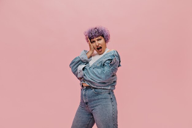 Free photo emotional adult woman with curly short purple hair is surprised on pink. modern lady in jeans clothes.