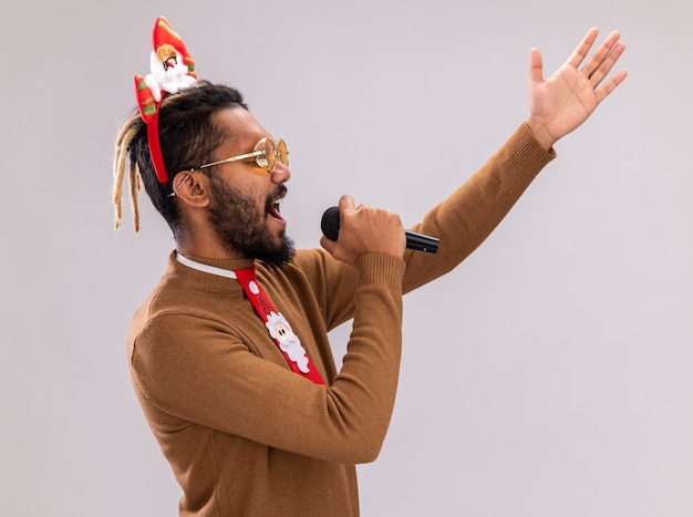 Free photo emotional african american man in brown sweater and santa rim on head with funny red tie shouting to microphone with raised arm standing over white background