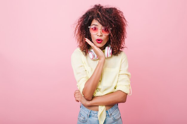 Emotional african woman wears stylish accessories posing in front of pink wall. Stunning lovable mulatto female model in jeans and sunglasses standing with surprised face expression.