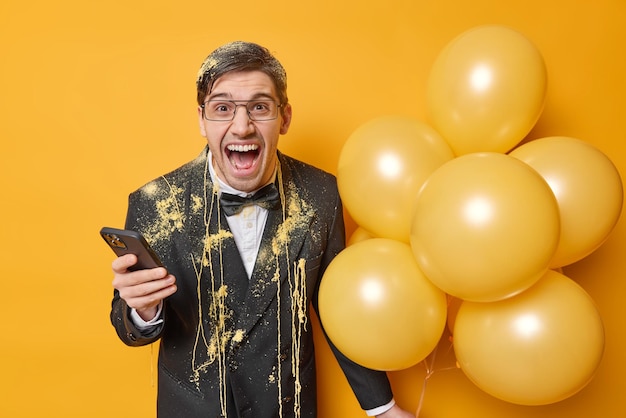 Emotional angry birthday man screams loudly dressed in elegant black suit holds mobile phone waits for call receives congratulations poses with inflated balloons isolated over yellow background