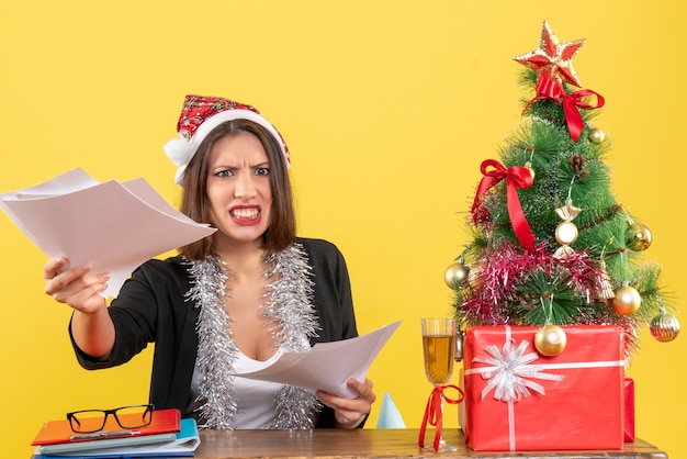 산타 클로스 모자와 새 해 장식 문서를 들고 크리스마스 트리가있는 테이블에 앉아 정장에 감정적 인 화가 비즈니스 아가씨
