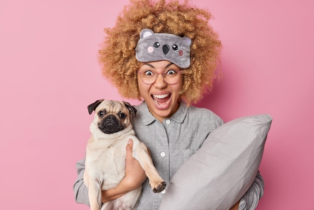 Emotional angry woman yells angrily wears pajama sleepmask on blindfold poses with pug dog holds pillow cannot fall asleep because of noise coming from neighbours isolated over pink background