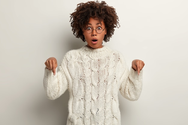 Free photo emotional attractive afro american woman points down with impressed shocked expression, wears optical glasses and jumper, models over white wall. advertisement concept. look downwards.