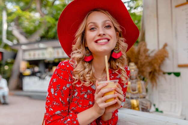 Emotional attractive stylish blond smiling woman in straw red hat and blouse summer fashion outfit drinking natural fruit cocktail smoothie