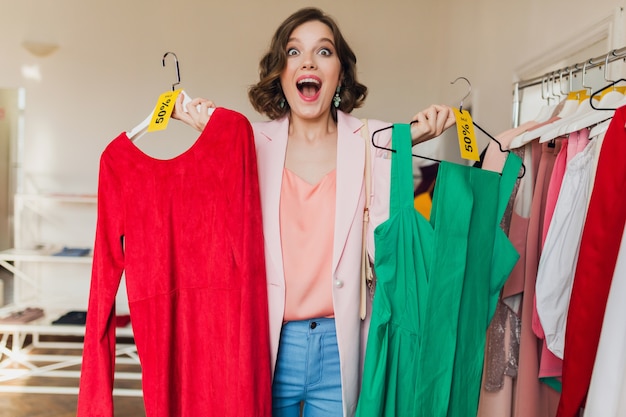 Emotional attractive woman holding colorful dresses on hanger in clothing store