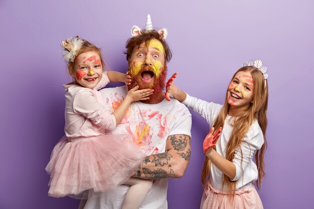 Emotional bearded busy father spends time with two naughty daughters who leave palm prints on his beard and clothes, learn how to paint, dressed in festive clothes, stand indoor. So colorful!