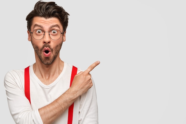 Emotional bearded male has surprised facial expression, astonished look, dressed in white shirt with red braces, points with index finger at upper right corner