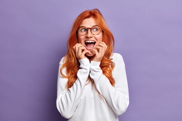 Emotional beautiful redhead woman grabs face looks gladfully aside keeps mouth widely opened wit amazement wears transparent glasses and white jumper reacts on unexpected surprise.