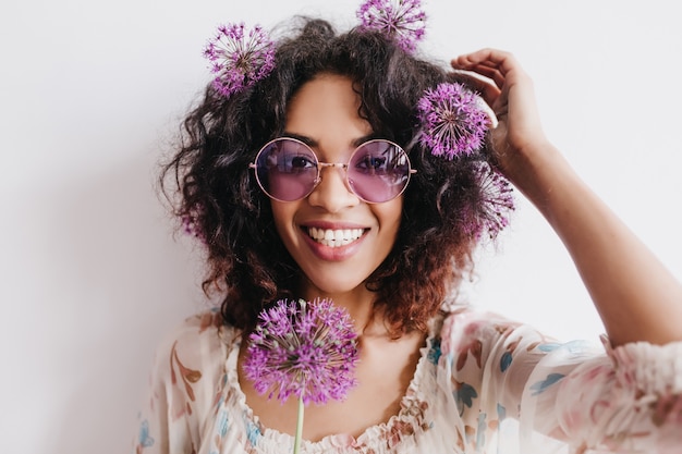 Эмоциональная черная женщина смеется, позируя с фиолетовыми цветами. Крытый снимок очаровательной африканской девушки с луком, развлекающейся.