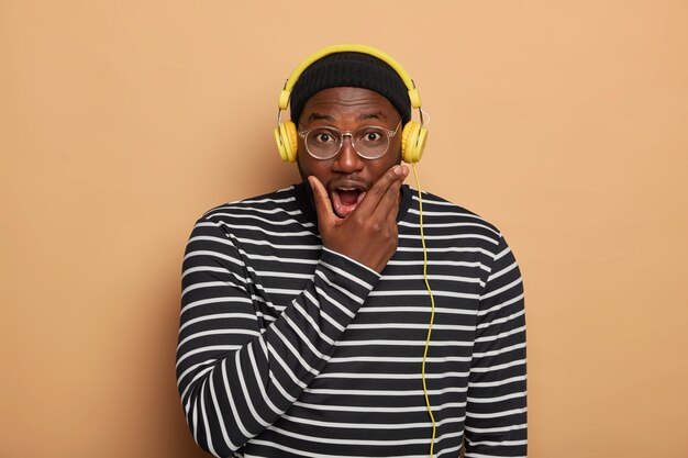 Emotional black young man holds chin, listens audio track in modern headphones