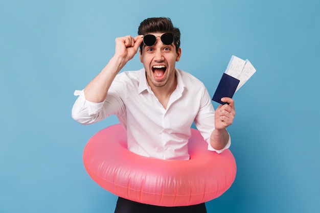 Emotional brown-eyed guy takes off his sunglasses and happily waving his passport and tickets. Man in white shirt posing with rubber ring against blue space.