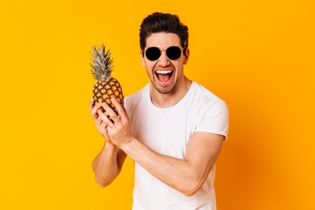 Emotional brunette man in sunglasses shouts and holds pineapple on orange background