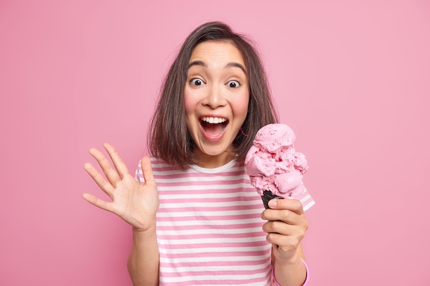 Emotional brunette woman exlaims loudly keeps palm raised eats delicious ice cream reacts on something unexpected has surpised joyfull look wears casual striped t shirt isolated over pink wall