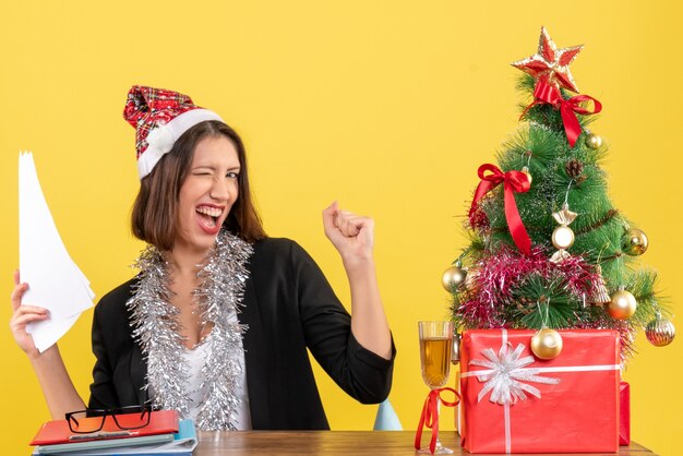 Emotional business lady in suit with santa claus hat and new year decorations holding documents and sitting at a table with a xsmas tree on it in the office