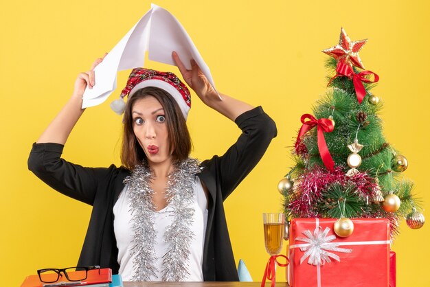 Emotional business lady in suit with santa claus hat and new year decorations raising documents on her head and sitting at a table with a xsmas tree on it in the office