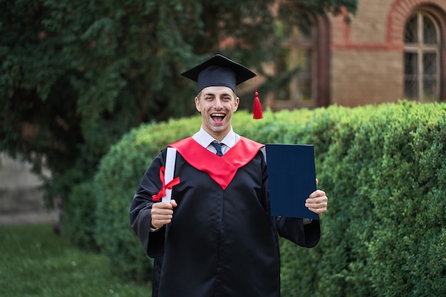 Emotional caucasian graduate in graduation robe and diploma in university campus.