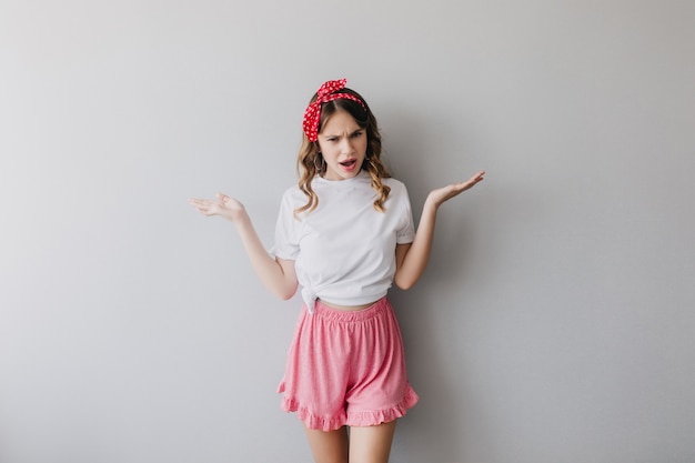 Free photo emotional caucasian lady with ribbon in hair waving hands. attractive woman in pink shorts standing.