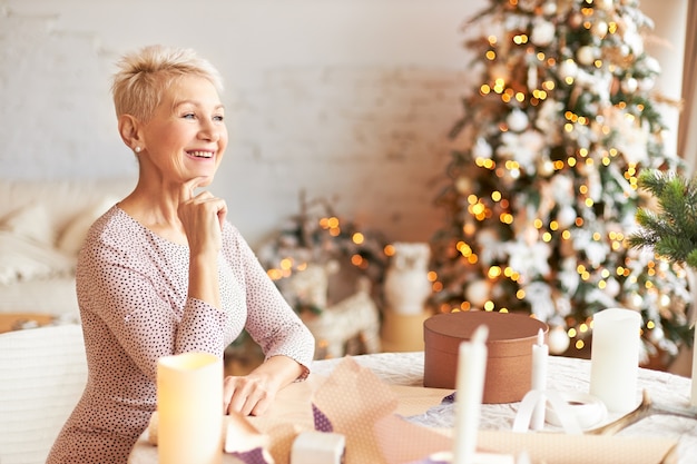 Emotional charming retired female with pixie hairstyle enjoying Christmas preparations wrapping presents in craft paper, having happy overjoyed facial expression, making gifts for family and friends