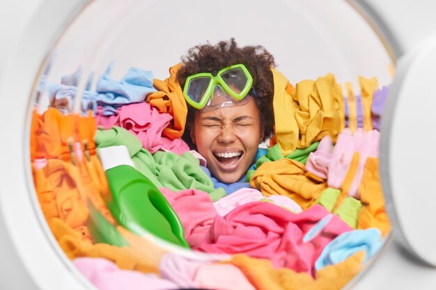 Emotional cheerful overjoyed Afro American housewife overloaded with housework has household duties sticks head in heap of colorful laundry wears snorkeling mask on forehead