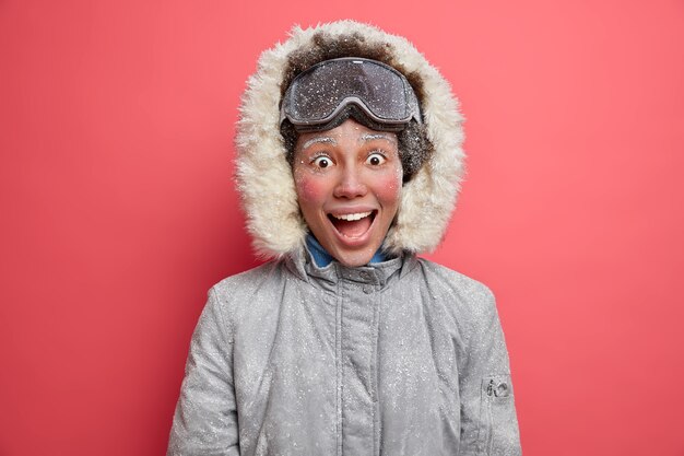 Emotional cheerful winter girl looks with overjoyed exprssion has red face covered by hoarfrost enjoys snowboarding during winter time wears warm jacket.