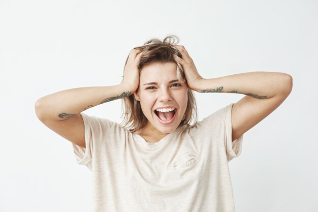 Emotional cheerful young tattooed girl smiling with opened mouth touching hair .