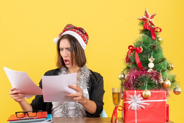 Emotional confused business lady in suit with santa claus hat and new year decorations looking at documents on her head and sitting at a table with a xsmas tree on it in the office