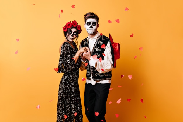 Эмоциональная пара, взявшись за руки, позирует для портрета в окружении конфетти сердца. нарядные костюмы парня и девушки дополняют свой необычный образ на хэллоуин.
